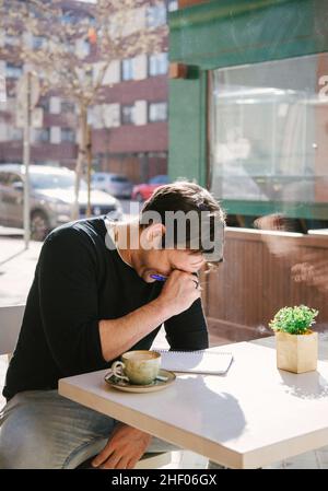 young man in a grey jacket writing and laughing something in note book while taking a coffee at the bar  Stock Photo