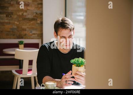 young man in a grey jacket holding a plant while taking a coffee at the bar  Stock Photo