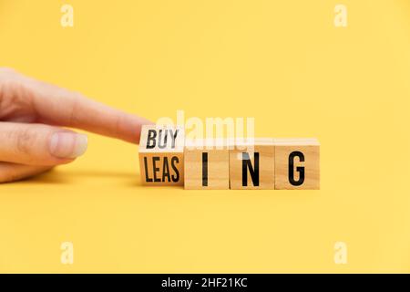 A woman's finger turns over a wooden cube, changing the inscription from buying to leasing. Yellow background with copy space. Close-up. Concept of mo Stock Photo