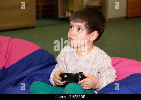 A handsome boy of 4 years old plays a game console, holds a joystick in his hands and looks away, a place for text Stock Photo