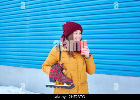smiling young woman with skates over her shoulder and a paper cup of coffee near the bright blue wall of the ice arena stadium. Stock Photo