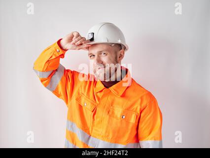 Smiling and looking at camera cute caucasian technician, engineer, supervisor or building contractor in white helmet and orange jacket correcting his working helmet. Building industry concept Stock Photo