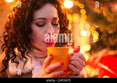 Young woman drinking hot drink from mug while standing against blurred decorated xmas tree outdoors during Christmas holidays, female with festive mak Stock Photo