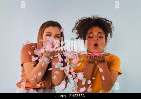 Two happy joyful multiracial girlfriends blowing confetti at camera and having fun while standing isolated over grey background, positive female frien Stock Photo