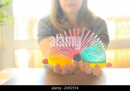Cropped shot of woman playing with plastic rainbow magic spring to relieve stress, middle-aged female holding bouncy stretchy toy while sitting at tab Stock Photo