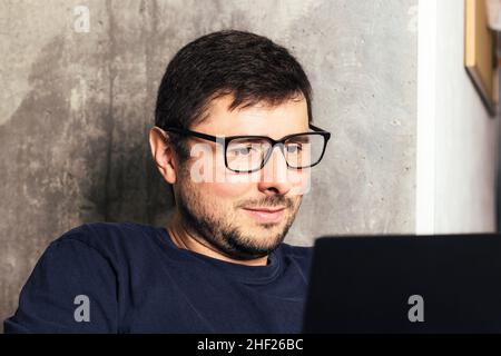 Handsome bearded man in glasses looking at his laptop screen against background of concrete wall in modern office Stock Photo