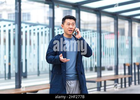 A man talking on the phone, focused and serious Asian at a public transport stop, a businessman near the office center Stock Photo