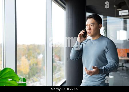 Serious and angry man talking on the phone in the office, pensive Asian at the window angry Stock Photo