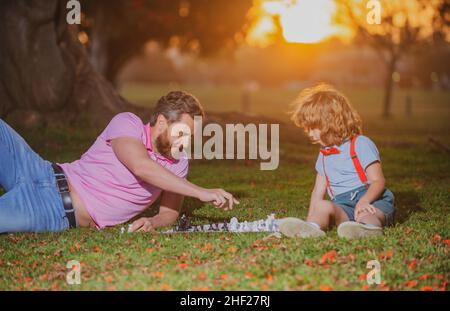 Father and son playing chess lying on grass at lawn park. Fathers Day, love family, parenthood, childhood concept. Active people having fun and Stock Photo