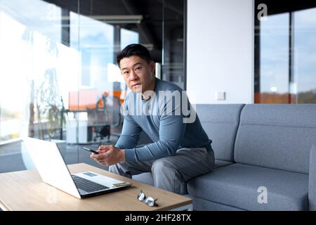 Serious and focused freelance businessman, in modern office, asian man kidnaps in camera, working on laptop, holding phone Stock Photo