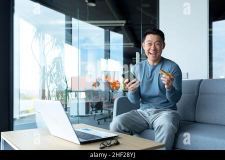 Happy asian male businessman holding credit card and phone, online shopping worker sitting in office Stock Photo