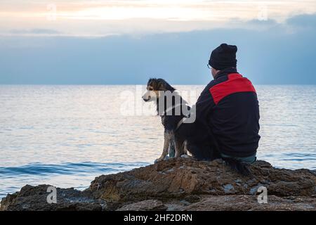 An unknown Caucasian man dressed in a black and red jacket, seen from his back, and his dog are sitting on a rock by the sea. The horizon is out of fo Stock Photo