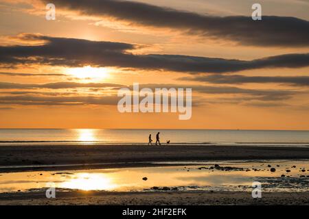 UK sunset beach landscape with young couple and pet dog silhouetted in the distance, walking on the shoreline sand. Stock Photo