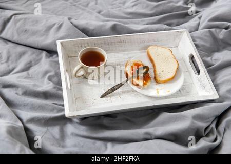A teapot, a cup of tea, two slices of toasted bread with apricot jam on a tray on the bedside table. Stock Photo