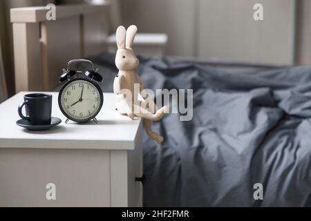 Image alarm clock with a black cup of coffee with figurine of wooden hare on a white bedside table in front of the bed with gray linens. The room is i Stock Photo