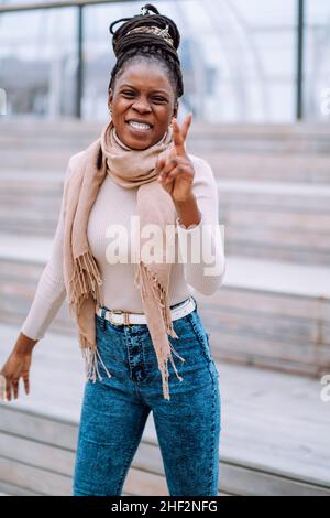 Afro-American woman in beige stole, roll-neck sweater and jeans with long afro braids gathered in bun showing V sign. Stock Photo