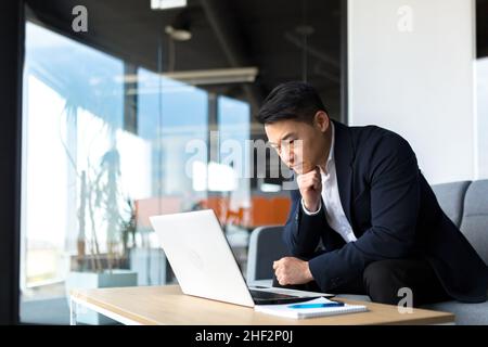 Portrait thoughtful Asian businessman working on a laptop computer at a modern office desk. Confident Focused pensive man in formal suit indoors. thin Stock Photo
