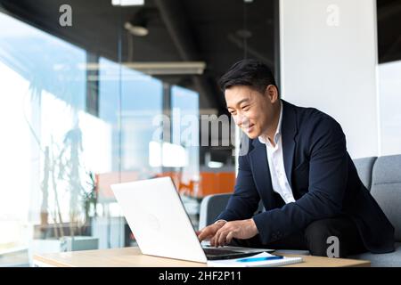 Senior Asian business man working on laptop online, smiling and rejoicing, happy boss business owner working in office Stock Photo