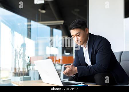Portrait thoughtful Asian businessman working on a laptop computer at a modern office desk. Confident Focused pensive man in formal suit indoors. thin Stock Photo