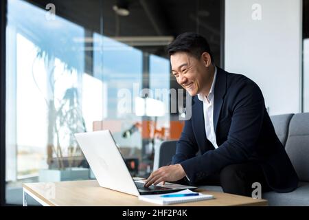 Senior Asian business man working on laptop online, smiling and rejoicing, happy boss business owner working in office Stock Photo
