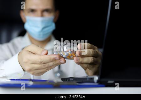 Bottle of capsules in hands of doctor in medical mask sitting at laptop in office. Concept of prescription, medication in pills, antibiotic or vitamin Stock Photo