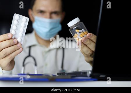 Pills and capsules in hand of doctor in medical mask sitting at laptop in office. Concept of prescription, choice of medicines, antibiotics or vitamin Stock Photo