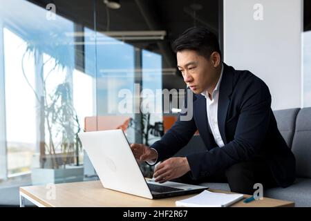 Concentrated male businessman talking to colleagues on video call using laptop sitting in office, asian man working online Stock Photo