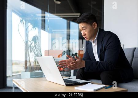 Concentrated male businessman talking to colleagues on video call using laptop sitting in office, asian man working online Stock Photo