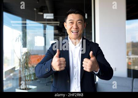 Asian happy man looking at camera showing thumbs up and ok sign, businessman smiling in modern office Stock Photo