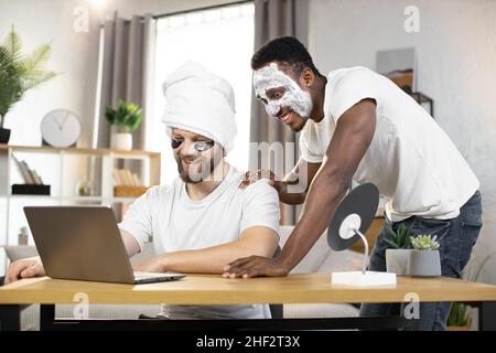 Attractive cheerful young multiracial two male friends with eye-patches and facial mask laughing when watching funny videos or photos on computer at home. Men skin care at home Stock Photo