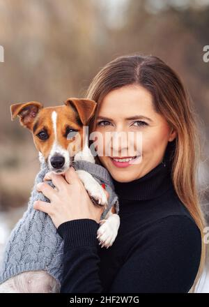 Young woman holding her Jack Russell terrier dog wearing warm winter clothing on hands Stock Photo