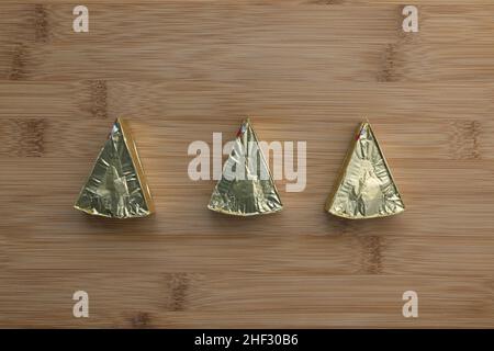 Pieces of processed cream cheese spread on wooden cutting board. Three triangles in gold foil. Minimalist background. Close-up, top view, copy space Stock Photo