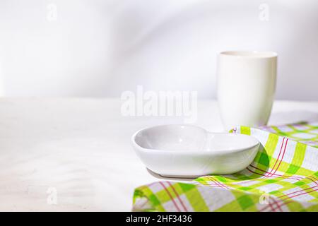 White ceramic heart shaped bowl with cup, the empty dishes are set for breakfast on a white stone kitchen surface, close-up, selective focus Stock Photo