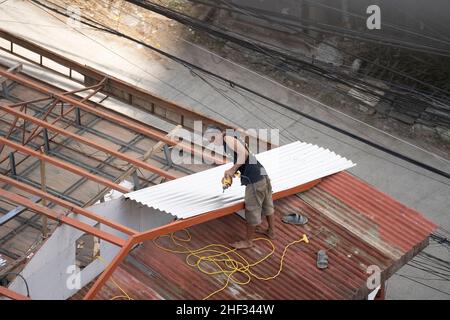 A man fixing galvanised sheets to a roof not wearing any shoes and using an electric drill. In the aftermath of Typhoon Odette, ( International Typhoon Rai), Cebu City, Philippines Stock Photo