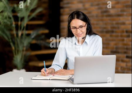 Smart focused attractive caucasian young woman wearing glasses and headphones, manager, support employee, online consultant, sit at laptop in office, records the appeal,conducts an online consultation Stock Photo