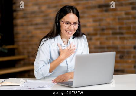 Satisfied happy sociable caucasian businesswoman wearing glasses and headset, support employee, online consultant, call center operator, using laptop, talking with client or employee, smiling Stock Photo