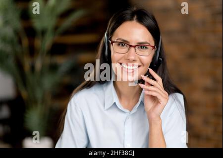 Pretty, positive caucasian brunette woman, wearing headset and glasses, freelance worker, call center employee, hot line operator, in formal shirt, looking at camera, smiles friendly Stock Photo