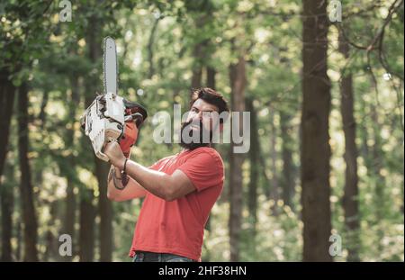 Lumberjack worker with chainsaw in the forest. Lumberjack worker walking in the forest with chainsaw. Deforestation. A handsome young man with a beard Stock Photo