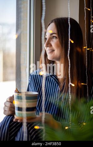 Happy domestic woman in pajamas holding cup of morning coffee standing near home window with garland Stock Photo