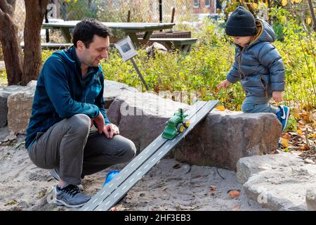 Father & son playing with toy tractor on ramp in city park sandbox Stock Photo