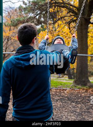 Father pushing two year old boy swinging on city park swing set; Corinthian Gardens; Philadelphia; Pennsylvania; USA Stock Photo