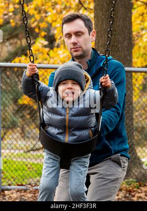 Father pushing two year old boy swinging on city park swing set; Corinthian Gardens; Philadelphia; Pennsylvania; USA Stock Photo