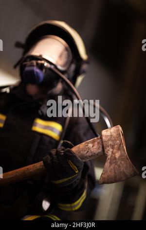 female firefighter portrait wearing full equipment, oxygen mask, and an axe. smoke and fire trucks in the background. Stock Photo