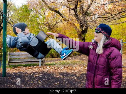 Senior grandmother pushing two year old boy swinging on city park swing set; Corinthian Gardens; Philadelphia; Pennsylvania; USA Stock Photo