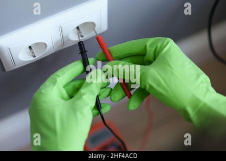 Hands in green gloves plug voltmeter into an outlet in wall Stock Photo