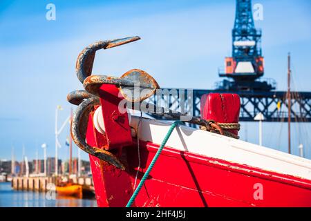 Rusty anchor on bow of old wooden cutter boat at city port background with huge gantry crane (copy space) Stock Photo