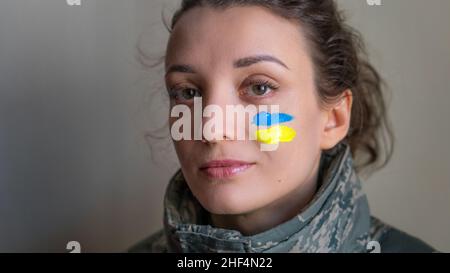 Indoor portrait of young girl with blue and yellow ukrainian flag on her cheek wearing military uniform, mandatory conscription in Ukraine, equality Stock Photo