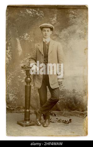 Original Edwardian era studio portrait photograph (cut down from a mount) of a handsome very confident looking older teenage boy / young man, casually holding a cigarette. A bit of a character. He is wearing middle class attire of a Town or Sack Suit, with a waistcoat from which can be seen a watch chain or 'fob', (indicating his status above working class) close-fit trousers. He wears a flat cap. dated 1910, U.K. Stock Photo