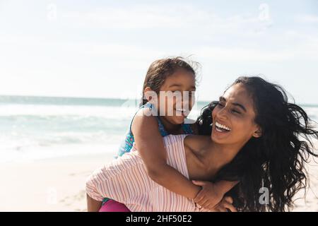 Cheerful biracial woman giving piggyback ride to daughter at beach against sky with copy space Stock Photo