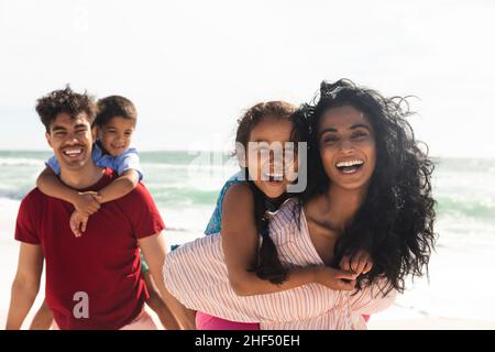 Portrait of happy multiracial parents giving piggyback rides to kids at beach with copy space Stock Photo